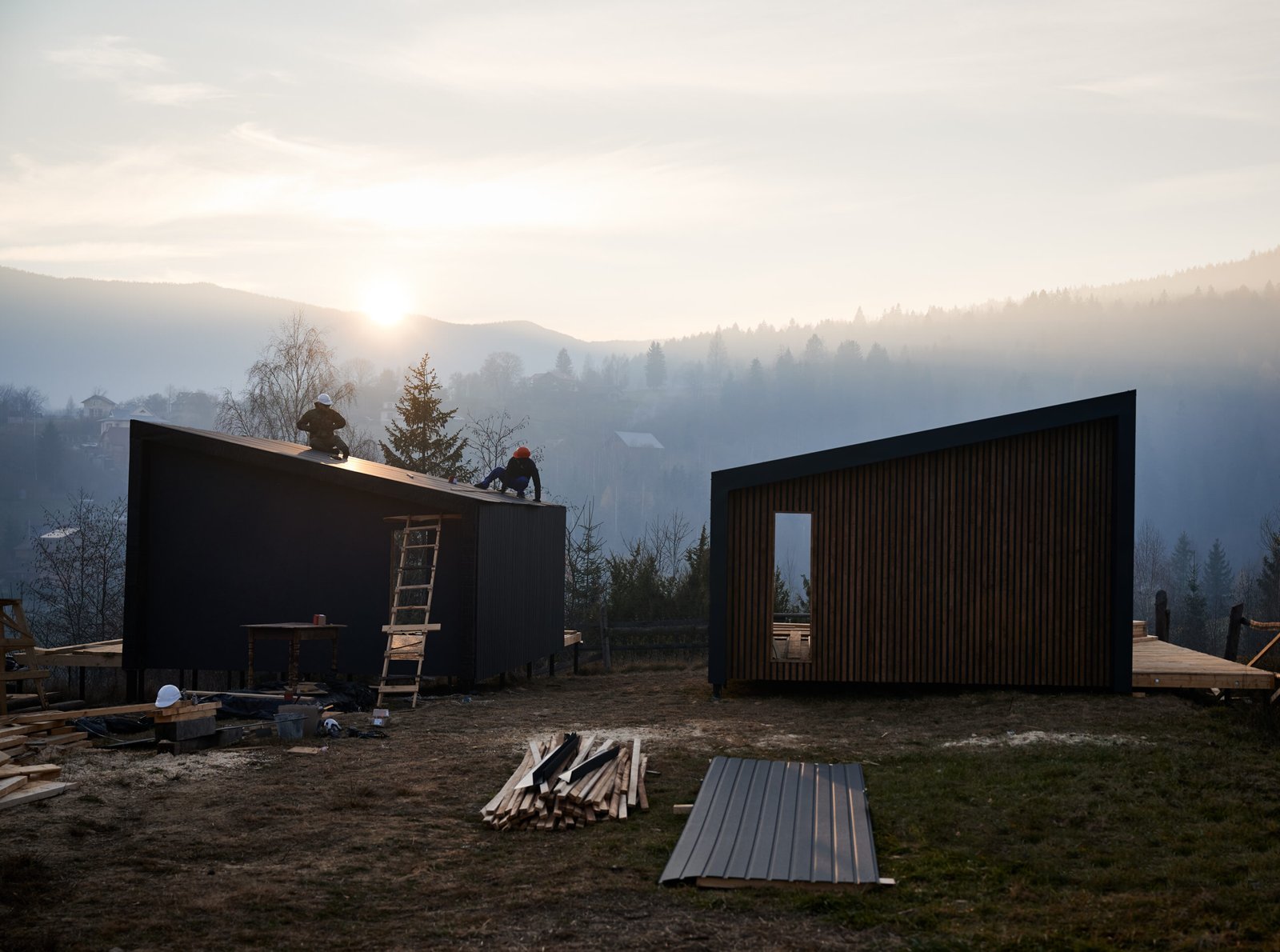 Male builders installing black corrugated iron sheet used as facade of future cottage at sunset. Men workers building wooden frame house. Carpentry and construction concept.